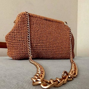 Peut inclure: Un sac à main marron en crochet avec une chaîne dorée. Le sac a une forme rectangulaire avec un dessus arrondi et une surface texturée. La chaîne dorée est épaisse et ajoute une touche d'élégance. Le sac est posé sur une surface grise.