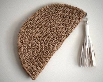 May include: A woven, half-moon shaped clutch in a natural tan color. The clutch has a white leather tassel attached to a silver-tone clasp. The texture of the clutch is visible, with a repeating pattern of curved lines.