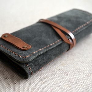 May include: A dark gray suede roll-up case with brown leather accents. The case is secured with a leather strap and features decorative stitching. The case has a small brown leather tab with two rivets.