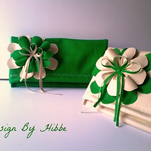 May include: Two decorative clutches, one green and one cream, each adorned with a large flower. The flowers feature green and cream petals with ribbon accents. The text "Design By Hibbe" is written in green cursive.
