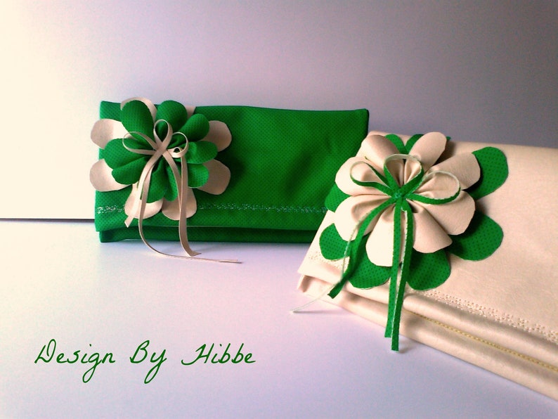 May include: Two decorative clutches, one green and one cream, each adorned with a large flower. The flowers feature green and cream petals with ribbon accents. The text "Design By Hibbe" is written in green cursive.