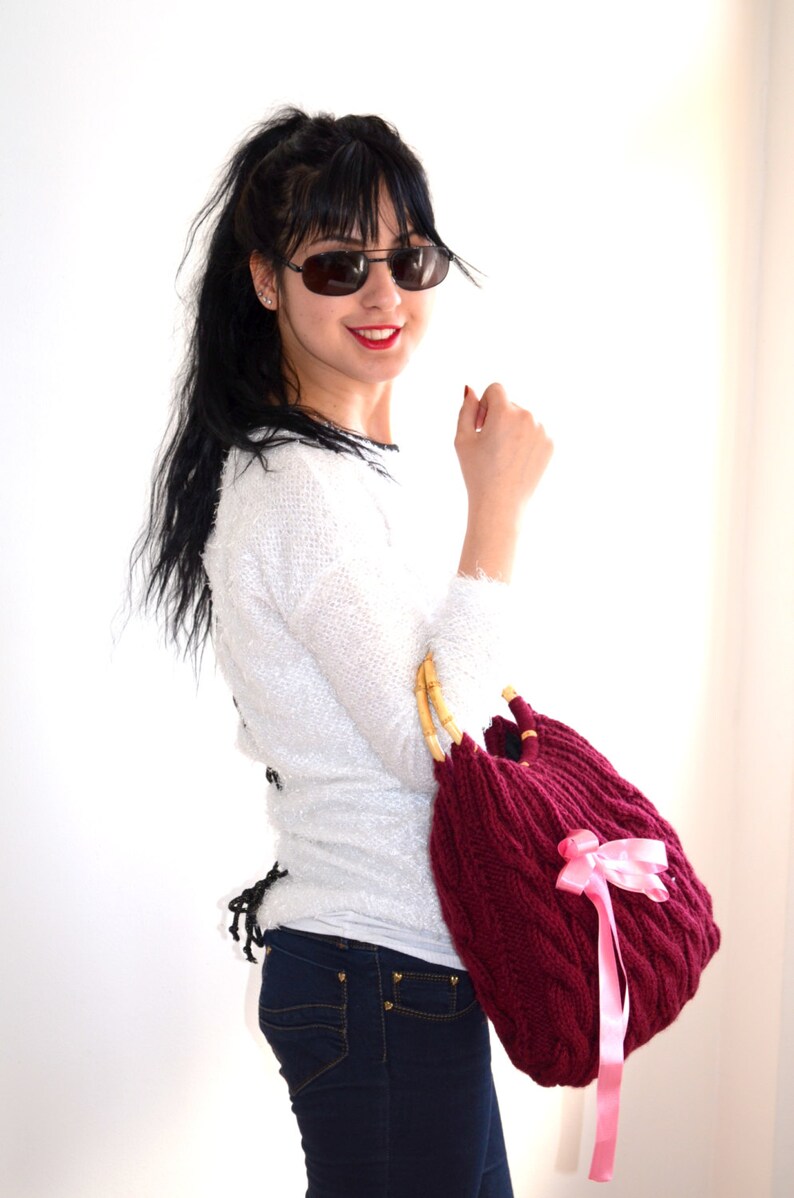 May include: A burgundy knitted handbag with bamboo handles and a pink ribbon bow. The bag has a cable knit design and is held by a person wearing sunglasses, a white sweater, and blue jeans.