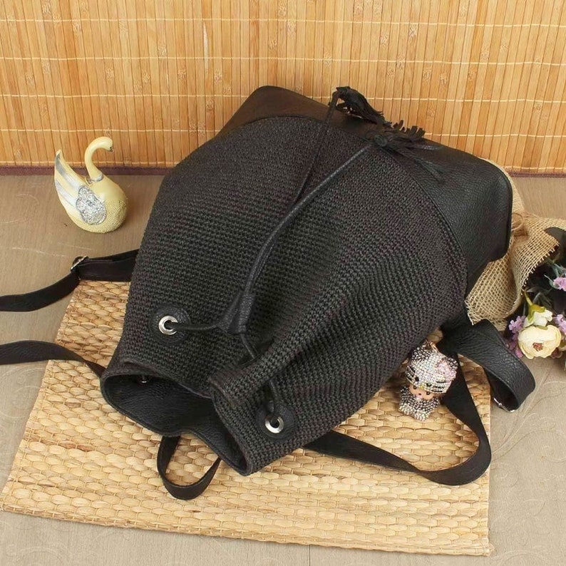 May include: Black backpack with a woven texture and leather accents. The drawstring closure and adjustable straps add functionality. A small tassel detail and silver-tone grommets enhance the design. The bag is set on a woven mat.