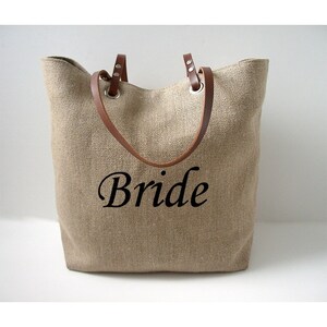 May include: A beige burlap tote bag with brown leather handles and the word "Bride" in black script. The bag has a wide opening and is designed for carrying items.