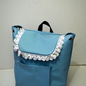 May include: A light blue backpack with a flap and a front pocket. The flap is trimmed with white lace. The bag has a black handle and metal eyelets.