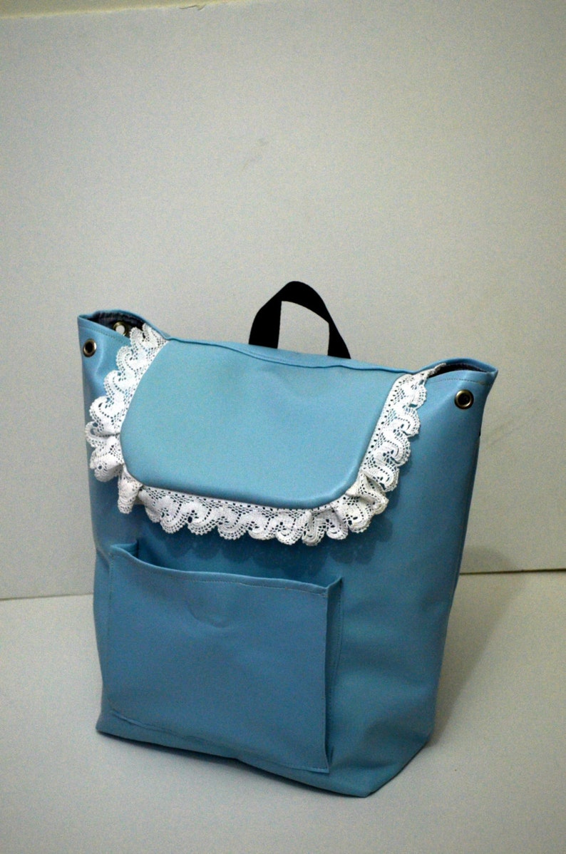 May include: A light blue backpack with a flap and a front pocket. The flap is trimmed with white lace. The bag has a black handle and metal eyelets.