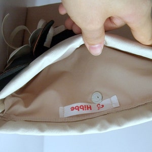 May include: A cream-colored clutch with a magnetic snap closure. The interior is a light tan color. A label with the word "Hibbe" in red is visible inside the clutch. The clutch is partially open, revealing a black and white floral design.
