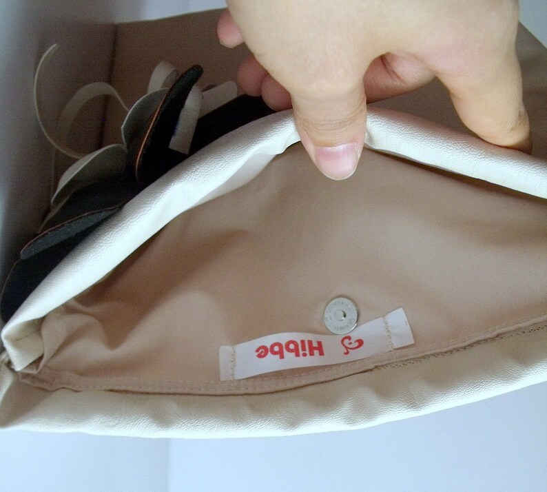 May include: A cream-colored clutch with a magnetic snap closure. The interior is a light tan color. A label with the word "Hibbe" in red is visible inside the clutch. The clutch is partially open, revealing a black and white floral design.