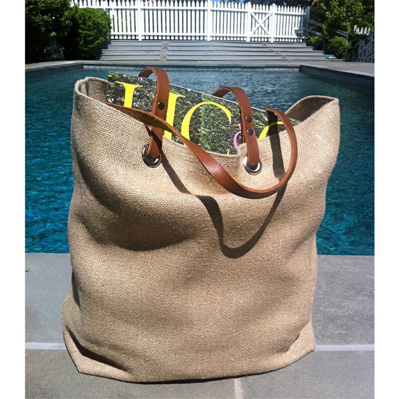 May include: A large, open-top tote bag made of tan burlap. The bag has brown leather handles attached with silver grommets. Magazines are visible inside the bag. The background features a swimming pool and a white picket fence.