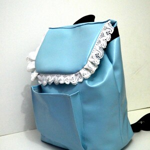 May include: A light blue backpack with a flap closure and a front pocket. The flap and edges are trimmed with white lace. The backpack has a black handle.