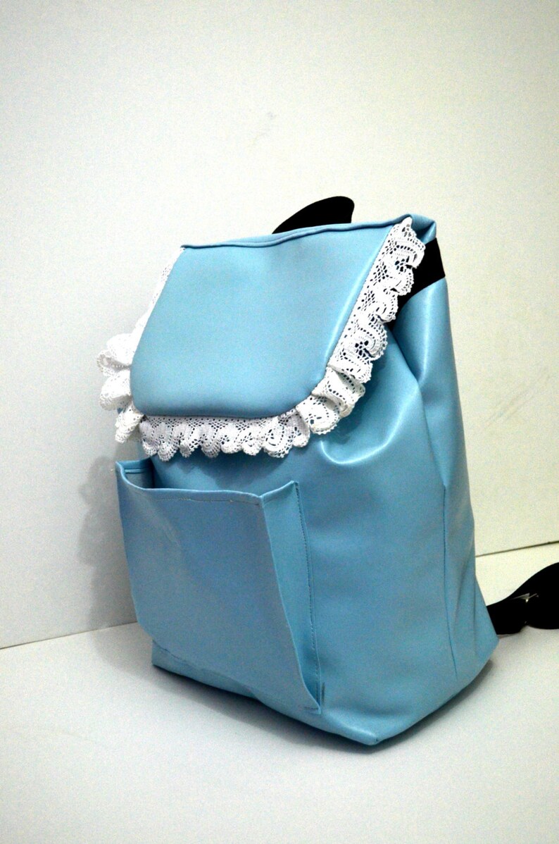 May include: A light blue backpack with a flap closure and a front pocket. The flap and edges are trimmed with white lace. The backpack has a black handle.