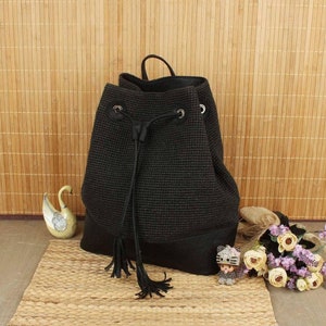 May include: A black drawstring backpack with a textured weave and a leather-like bottom. The bag has a top handle and a drawstring closure with tassels. The background features a woven mat and bamboo blinds.