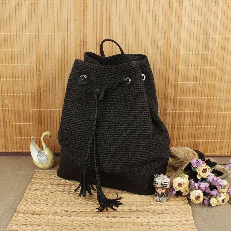 May include: A black drawstring backpack with a textured weave and a leather-like bottom. The bag has a top handle and a drawstring closure with tassels. The background features a woven mat and bamboo blinds.