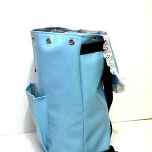 May include: A light blue backpack with a black strap and a small side pocket. The top of the bag has metal grommets and a black interior. A white lace trim is visible on the side.