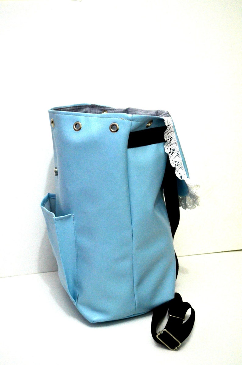 May include: A light blue backpack with a black strap and a small side pocket. The top of the bag has metal grommets and a black interior. A white lace trim is visible on the side.