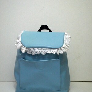 May include: A light blue backpack with a front pocket and a flap closure. The flap is trimmed with white lace. The backpack has a black handle at the top.