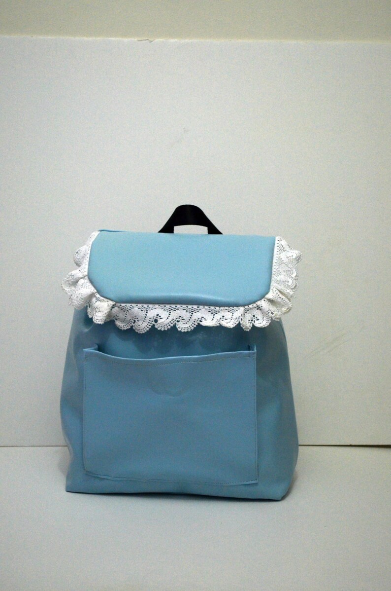 May include: A light blue backpack with a front pocket and a flap closure. The flap is trimmed with white lace. The backpack has a black handle at the top.
