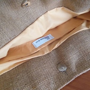 May include: Open view of a burlap tote bag with a light tan interior lining. The bag has a magnetic snap closure and a small, rectangular pocket inside. The interior lining is a light peach color. The bag is made of a woven, natural fiber material.