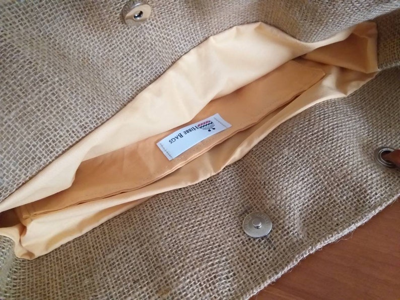 May include: Open view of a burlap tote bag with a light tan interior lining. The bag has a magnetic snap closure and a small, rectangular pocket inside. The interior lining is a light peach color. The bag is made of a woven, natural fiber material.