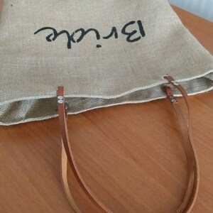 May include: A natural burlap tote bag with the word "Bride" in black script. The bag features brown leather handles and is designed for carrying items. The bag is sitting on a wooden surface.