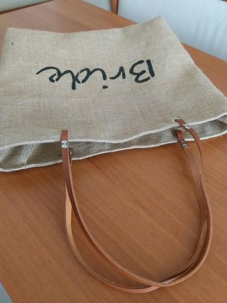 May include: A natural burlap tote bag with the word "Bride" in black script. The bag features brown leather handles and is designed for carrying items. The bag is sitting on a wooden surface.