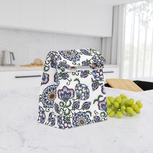 May include: White lunch bag with a colorful floral pattern in blue, red, and green. The bag is sitting on a white countertop in a kitchen setting. A bunch of green grapes is next to the bag.