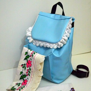 May include: A light blue backpack with a black handle and strap, trimmed with white lace. Two embroidered cloths with floral patterns in pink, green, and purple are displayed in front of the backpack.