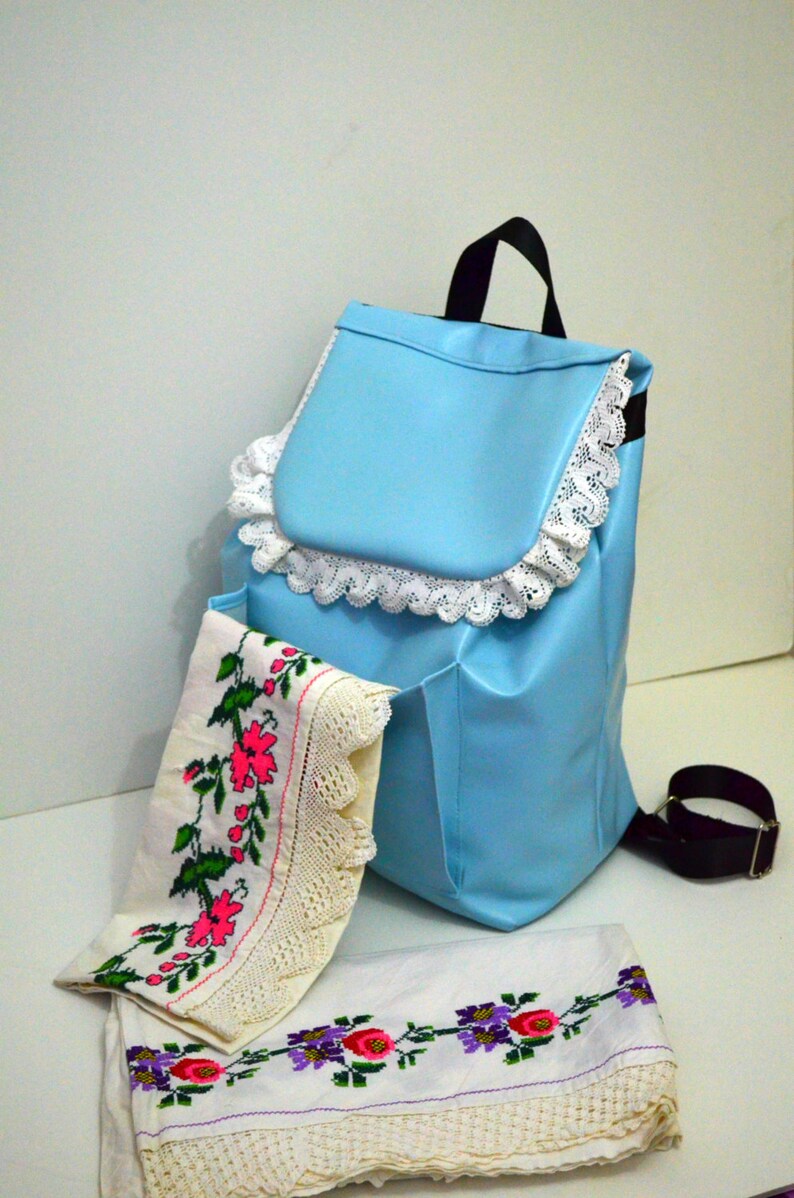 May include: A light blue backpack with a black handle and strap, trimmed with white lace. Two embroidered cloths with floral patterns in pink, green, and purple are displayed in front of the backpack.
