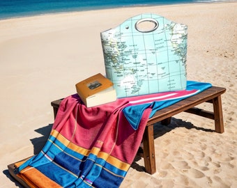 May include: A beach scene featuring a wooden bench with a red and blue striped beach towel, a book, and a tote bag with a world map design. The tote bag has a silver handle. The background shows a sandy beach and the ocean.