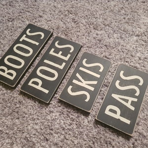 Può includere: Quattro cartelli in legno nero con lettere bianche. I cartelli recitano "BOOTS", "POLES", "SKIS" e "PASS".
