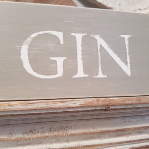 May include: A rustic wooden sign with a distressed white paint finish. The sign reads "GIN" in white lettering.