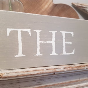 May include: A gray wooden sign with white painted lettering that reads "THE G".