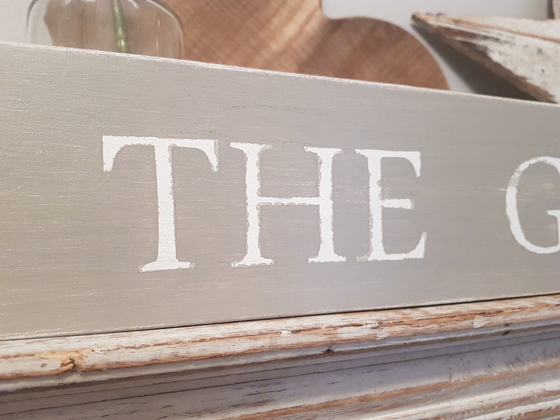May include: A gray wooden sign with white painted lettering that reads "THE G".