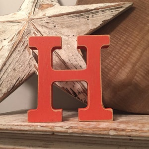 Puede incluir: Una vibrante letra "H" de madera naranja se encuentra sobre una estantería desgastada pintada de blanco. Una gran estrella blanca desgastada y un cuenco de madera marrón en el fondo crean una estética rústica de granja. La letra es un acento decorativo.