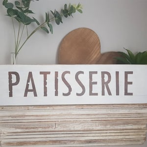 Può includere: Un cartello rettangolare in legno bianco con la scritta "PATISSERIE" in lettere marroni. Il cartello è esposto su una mensola bianca invecchiata, con un cuore di legno e piante verdi sullo sfondo. Decorazione per cucina o bar.