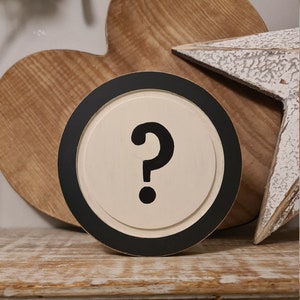 May include: A round wooden sign with a black border and a white background. The sign has a large black question mark on it.