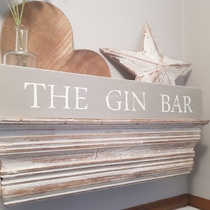 May include: A gray wooden sign with white lettering that reads "THE GIN BAR". The sign is mounted on a white wooden shelf with decorative trim.