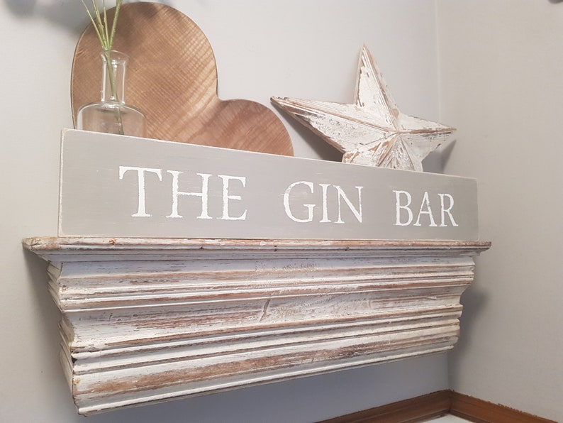 May include: A gray wooden sign with white lettering that reads "THE GIN BAR". The sign is mounted on a white wooden shelf with decorative trim.