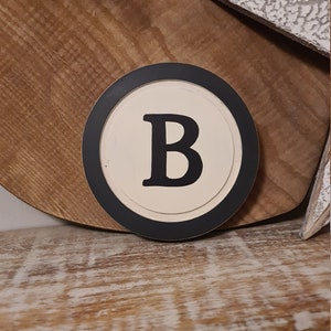 May include: A black and white wooden letter B with a black border. The letter is on a wooden surface.