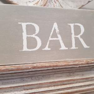 May include: A gray wooden sign with the word "BAR" painted in white on it. The sign has a distressed white and brown frame.