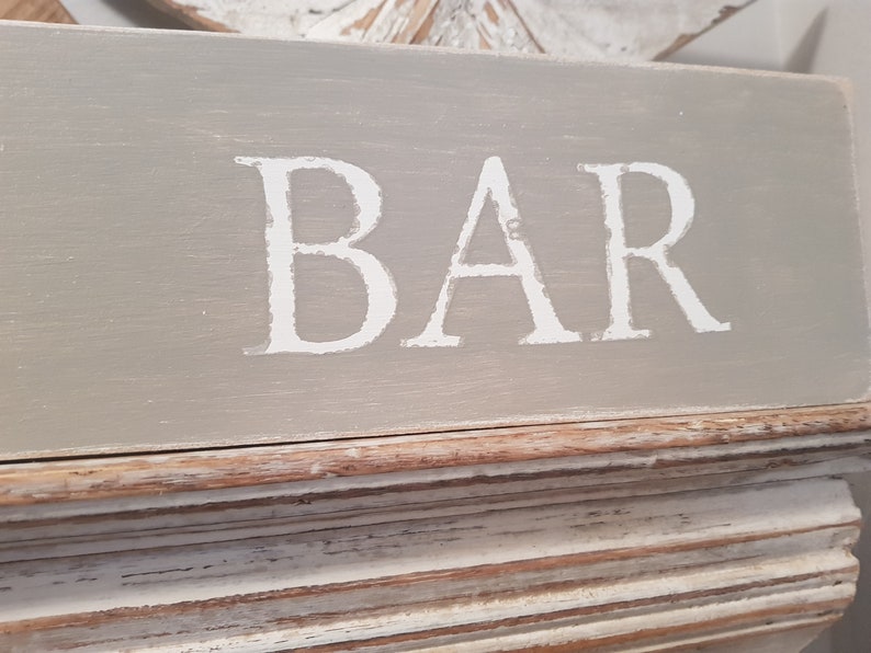 May include: A gray wooden sign with the word "BAR" painted in white on it. The sign has a distressed white and brown frame.