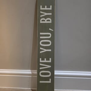 May include: A rectangular, olive green wooden sign with the words "LOVE YOU, BYE" in white, vertically aligned. The sign is leaning against a wall.