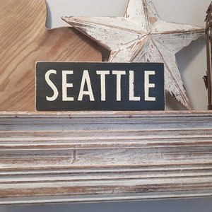 Puede incluir: Un letrero de madera negro con letras blancas que dice "SEATTLE". El letrero está sentado en un estante blanco con un corazón de madera y una estrella blanca a cada lado.