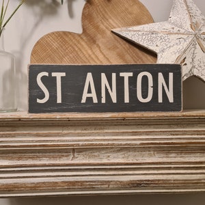 May include: A black wooden sign with white lettering that reads "ST ANTON" on a white shelf with a wooden heart and a white star.