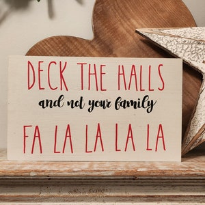 Könnte beinhalten: Ein weißes Holzschild mit rotem Text, auf dem "Deck the halls and not your family" und "Fa la la la la" steht.