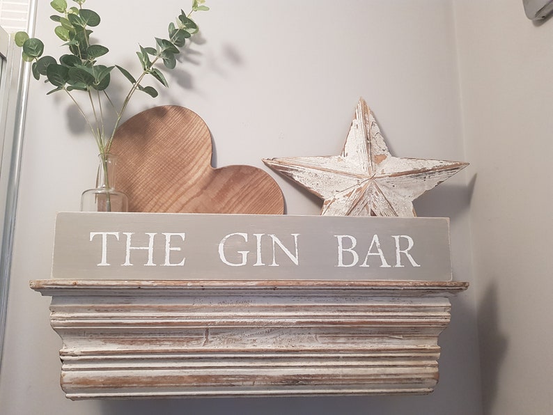 May include: A gray wooden sign with the text "THE GIN BAR" in white lettering. The sign is on a white shelf with a wooden heart and a white wooden star on top of it.