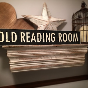 Puede incluir: Un letrero de madera negro con letras blancas que dice "OLD READING ROOM". El letrero está montado en un estante blanco con molduras decorativas. Hay una estrella blanca y una jaula de pájaros en el fondo.