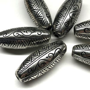 May include: Close-up of several silver-colored, oblong beads. Each bead features intricate etched designs, including geometric patterns and swirling lines. The beads are arranged on a white surface, showcasing their detailed craftsmanship.