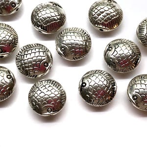 May include: Silver-colored, round beads with a fish design. Each bead has a detailed fish scale pattern and a small hole for stringing. The beads are scattered on a white surface.