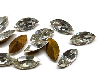 May include: A group of clear, faceted, marquise-shaped rhinestones, some with a gold backing, scattered on a white surface.